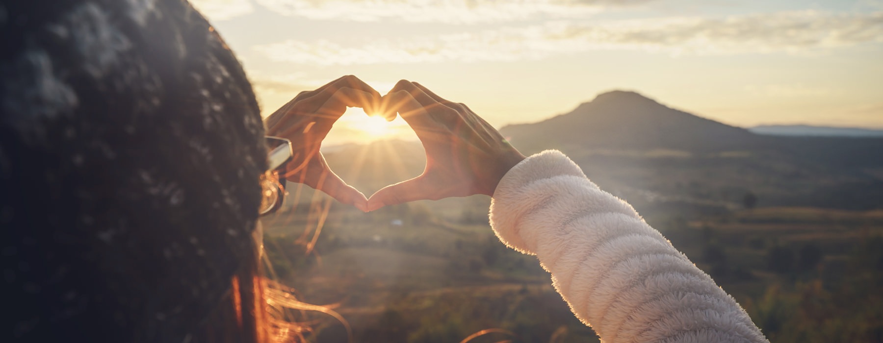 woman makes a heart shape around sunrise over mountains with her hands