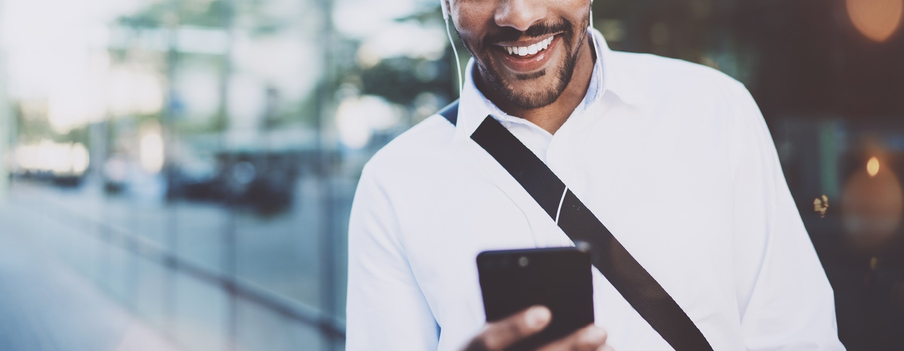 smiling man walks down city sidewalk as he looks at his phone